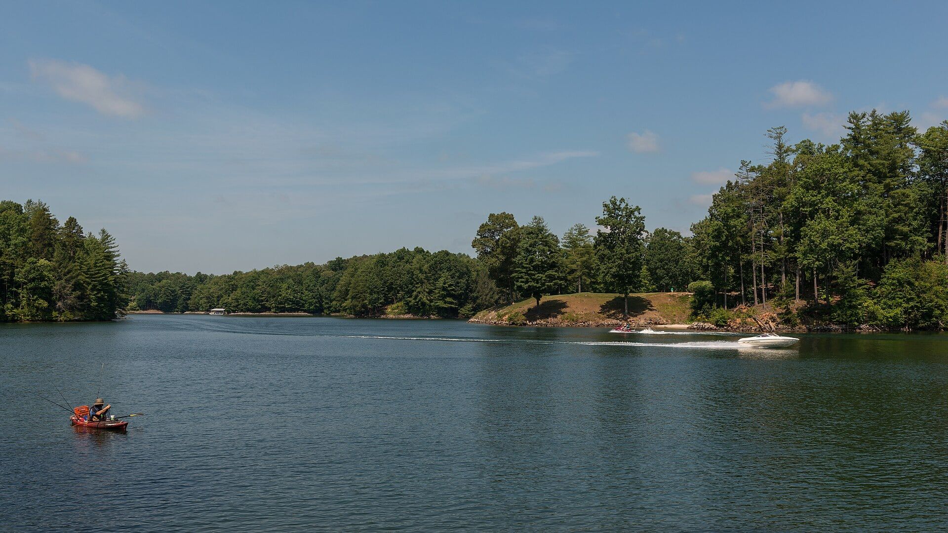 Lake Keowee on a summer day from Keowee-Toxaway State Park: a kayaker fishing, a boat's wake and a wooded shoreline.